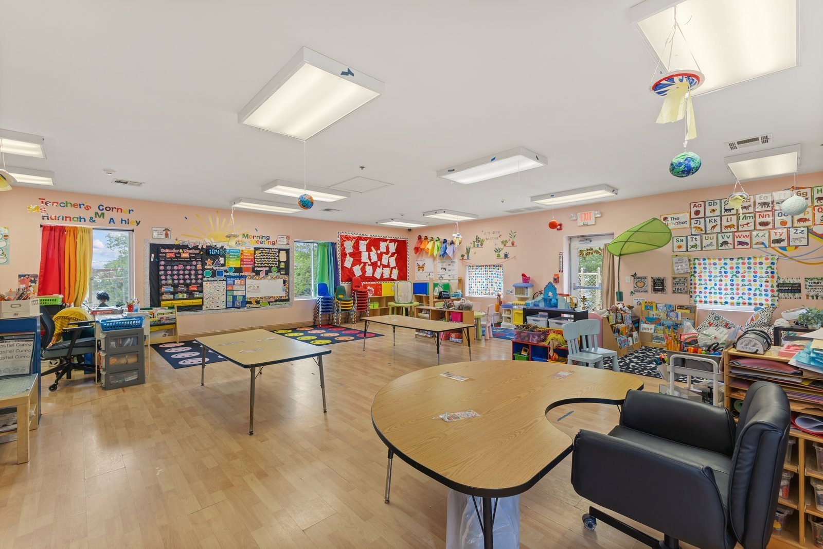 Wide view of the bright Sunny Days Pre-K classroom with child-size tables, a circle time rug, reading corner, alphabet wall and hanging planet mobiles.